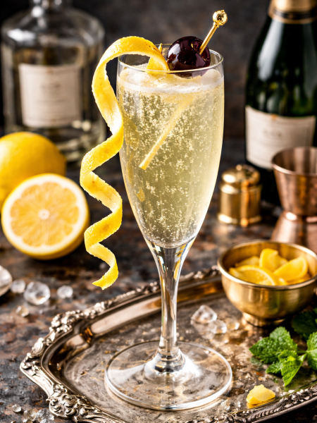 French 75 cocktail in champagne flute with lemon twist and cherry garnish on silver tray with fresh lemons and bar tools