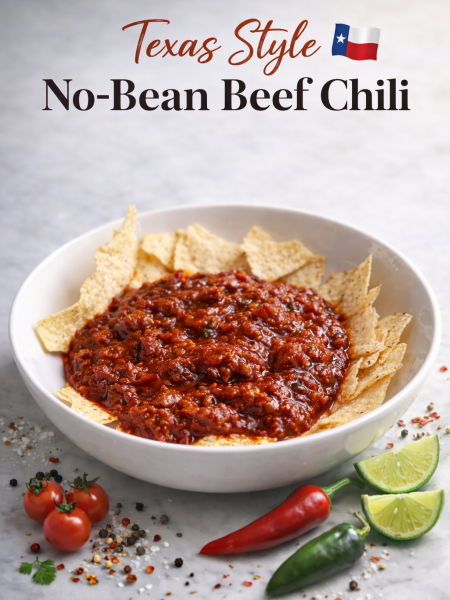 Texas-style no-bean beef chili in a rustic ceramic bowl, topped with shredded cheddar cheese, fresh jalapeño slices, and a dollop of sour cream, with a wooden spoon resting on the side