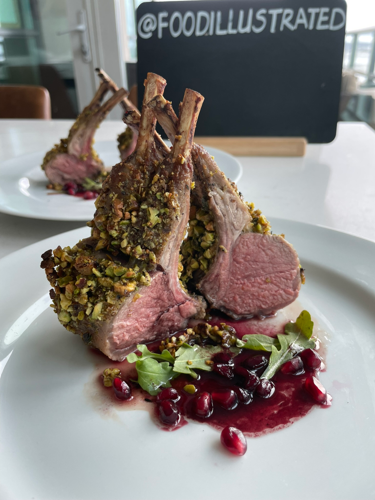 A elegant French bistro style pistachio crusted rack of lamb sliced into perfect pink chops, drizzled with a rich red pomegranate wine sauce on a white plate.
