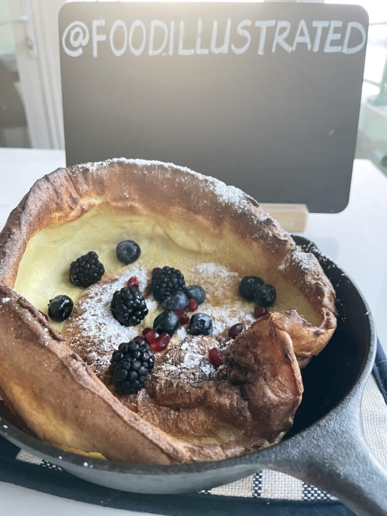Golden, dramatically puffed Dutch baby pancake in a black cast iron skillet, dusted with powdered sugar, surrounded by fresh strawberries and blueberries.