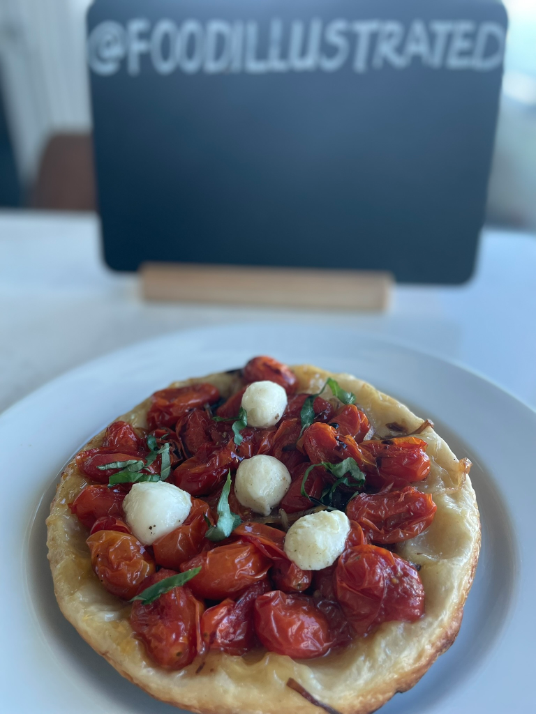 A golden brown Caramelized Tomato Tarte Tatin on a white platter revealing layers of jammy cherry tomatoes, caramelized onions, and olives upside down.